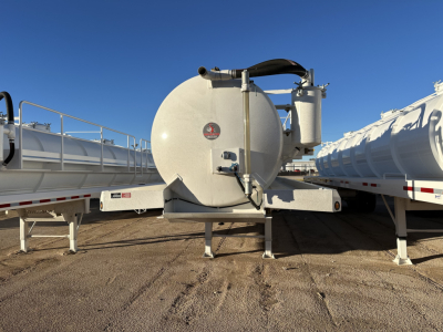  tank trailer with circular rear end and black hose, on a tri-axle chassis, with adjacent trailers on both sides and clear blue sky