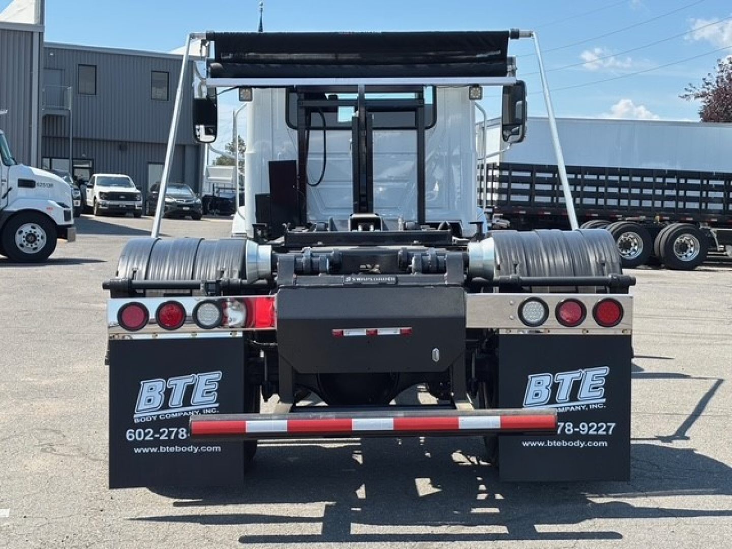 Rear view of a new 2026 Mack MD7 hooklift truck showing bumper with BTE logos, multiple taillights, chrome bumper guards, and a hydraulic/cable system for the hooklift.