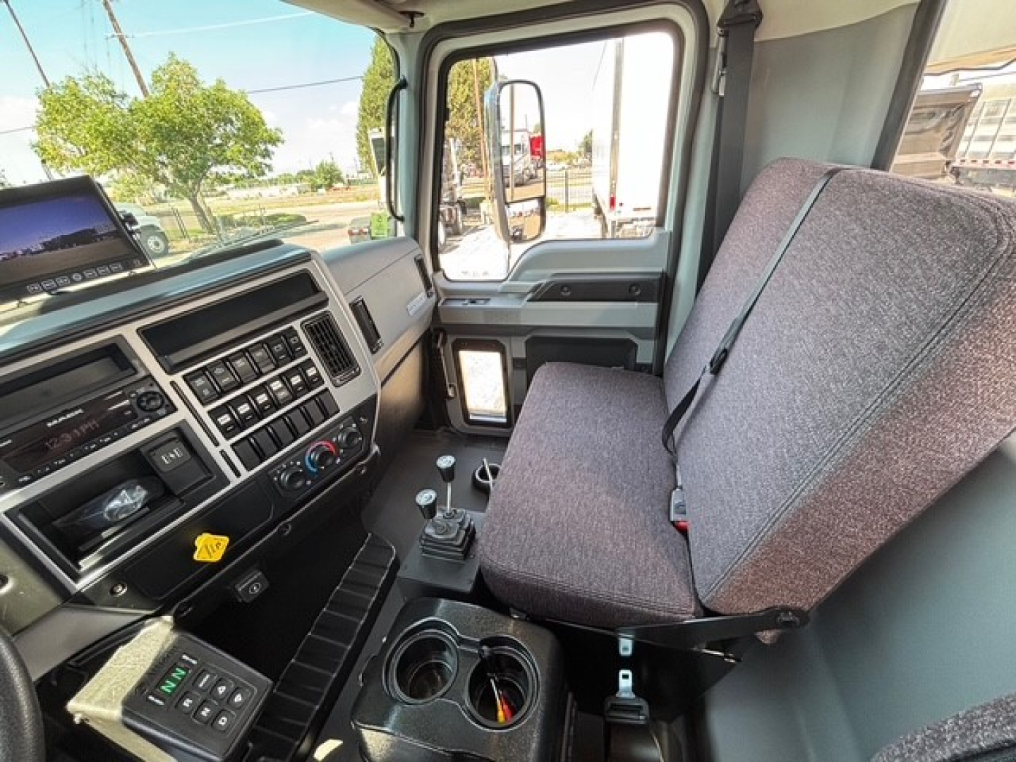 Cab interior of a new 2026 Mack MD7 hooklift truck showing driver seat, steering wheel, center console, gear shifter, and dashboard controls with window view.