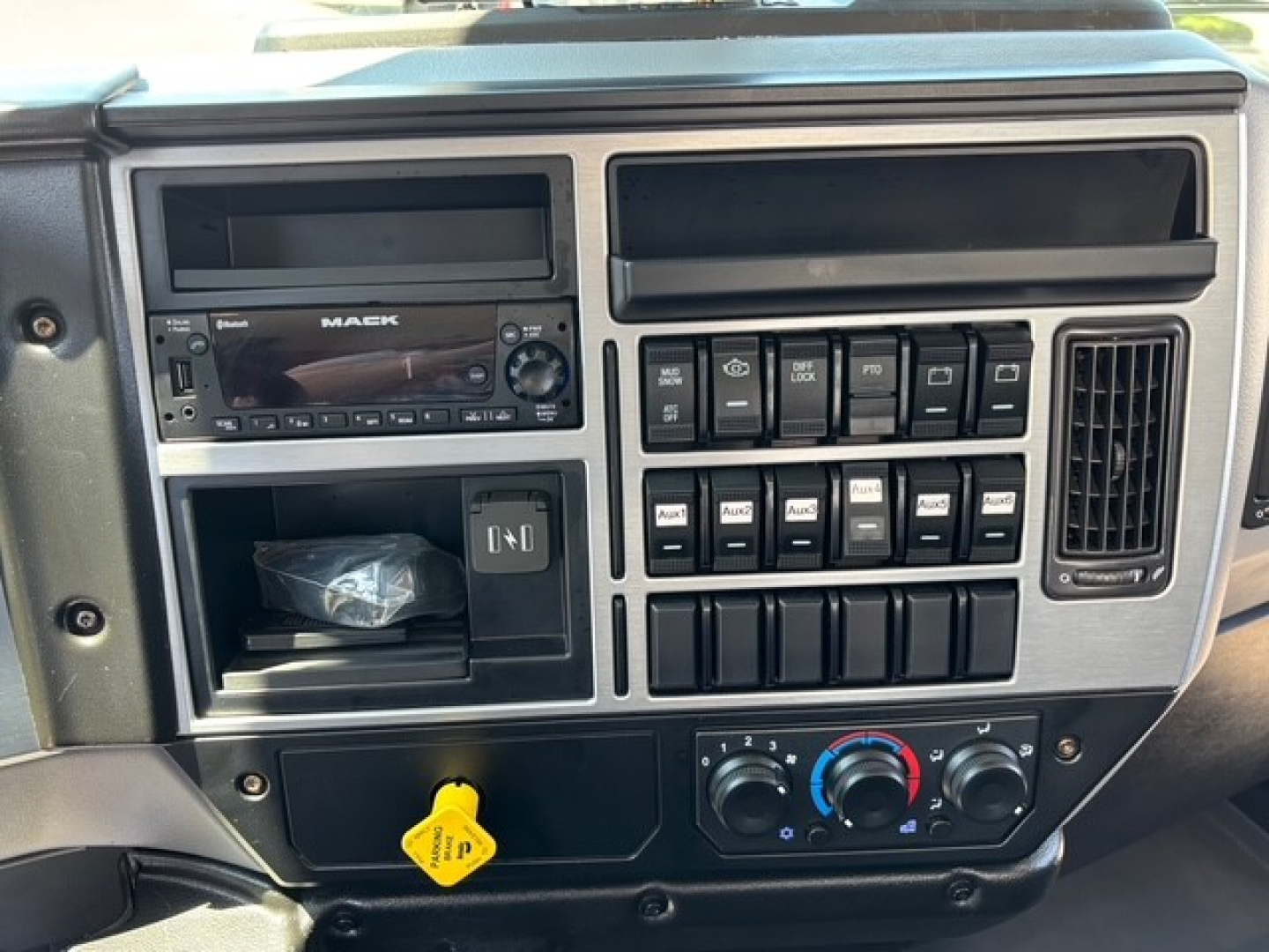 Mack MD7 dashboard/center console showing radio, control switches, auxiliary storage, and climate controls with yellow hood release sticker.