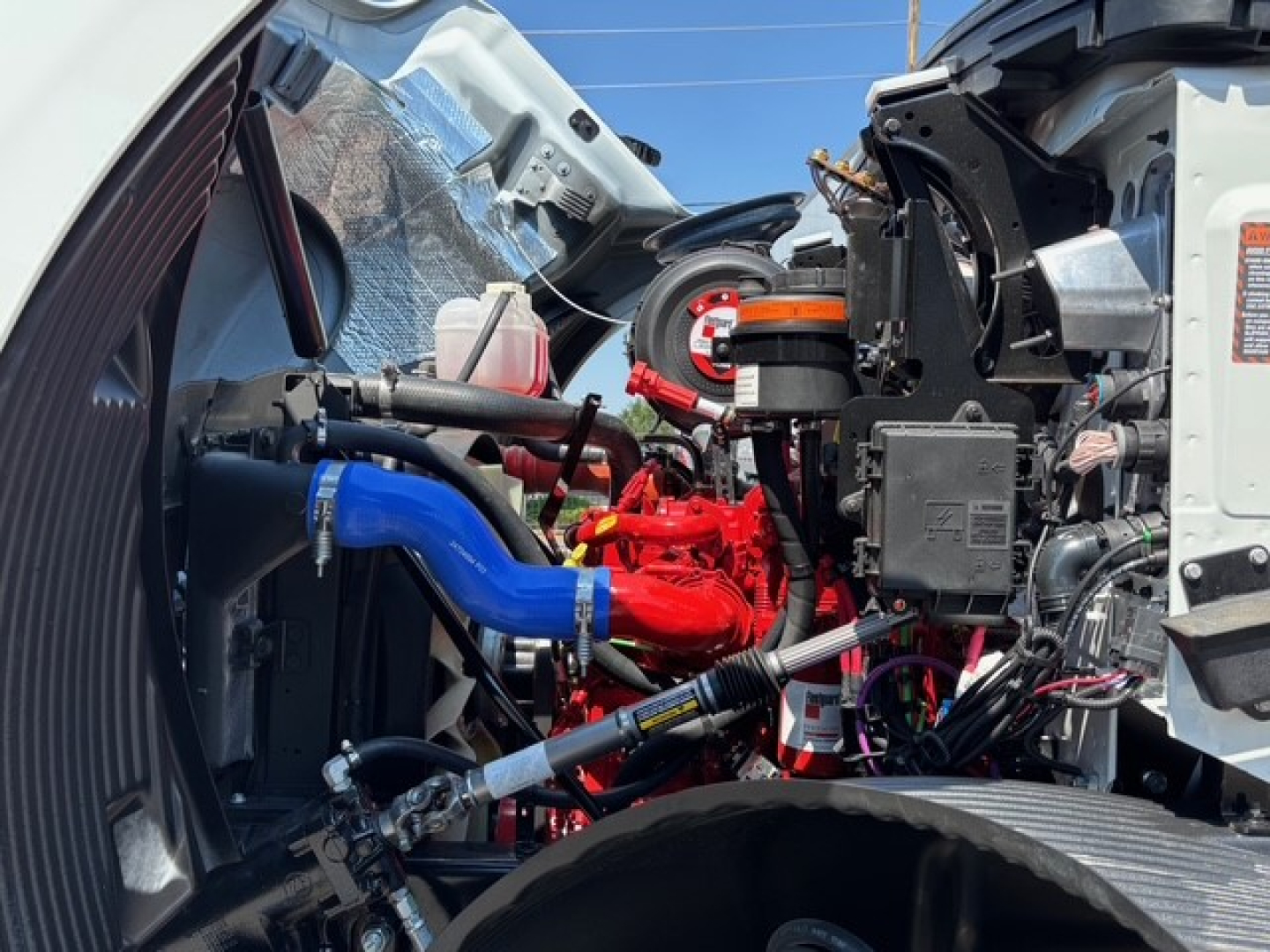 Engine bay of a 2026 Mack MD7 hooklift showing red engine components, blue hoses, and black electrical harnesses with exposed wiring.
