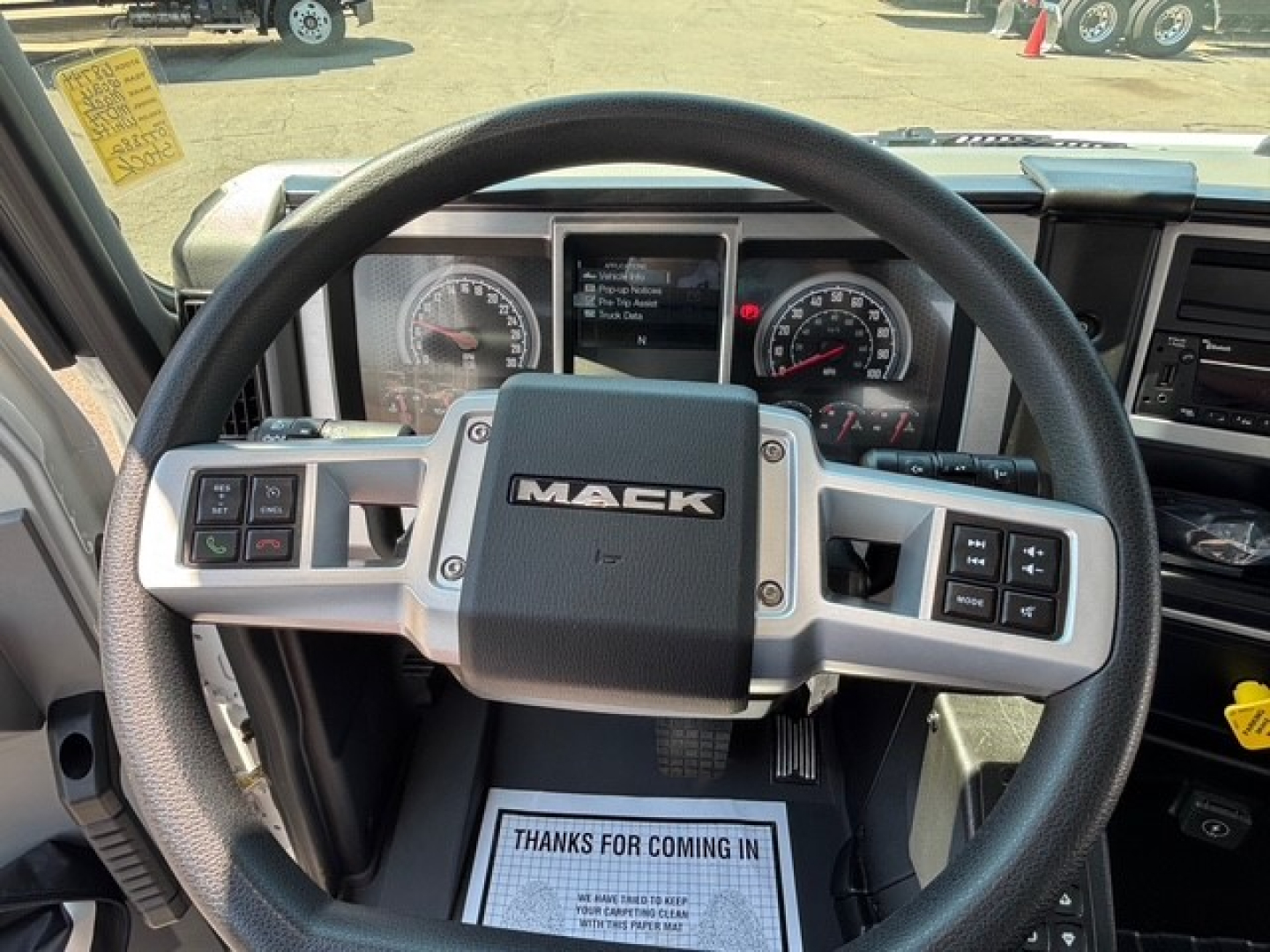 Steering wheel and instrument cluster of a Mack MD7 truck cab; visible dashboard gauges and controls with 'MACK' logo on center; windshield view of yard.