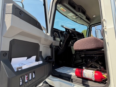 Cab interior of a 2026 Mack MD7 hooklift truck showing driver's seat, steering wheel, dashboard, open door with storage pocket, and a fire extinguisher on the floor.