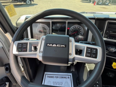 Steering wheel and instrument cluster of a Mack MD7 truck cab; visible dashboard gauges and controls with 'MACK' logo on center; windshield view of yard.