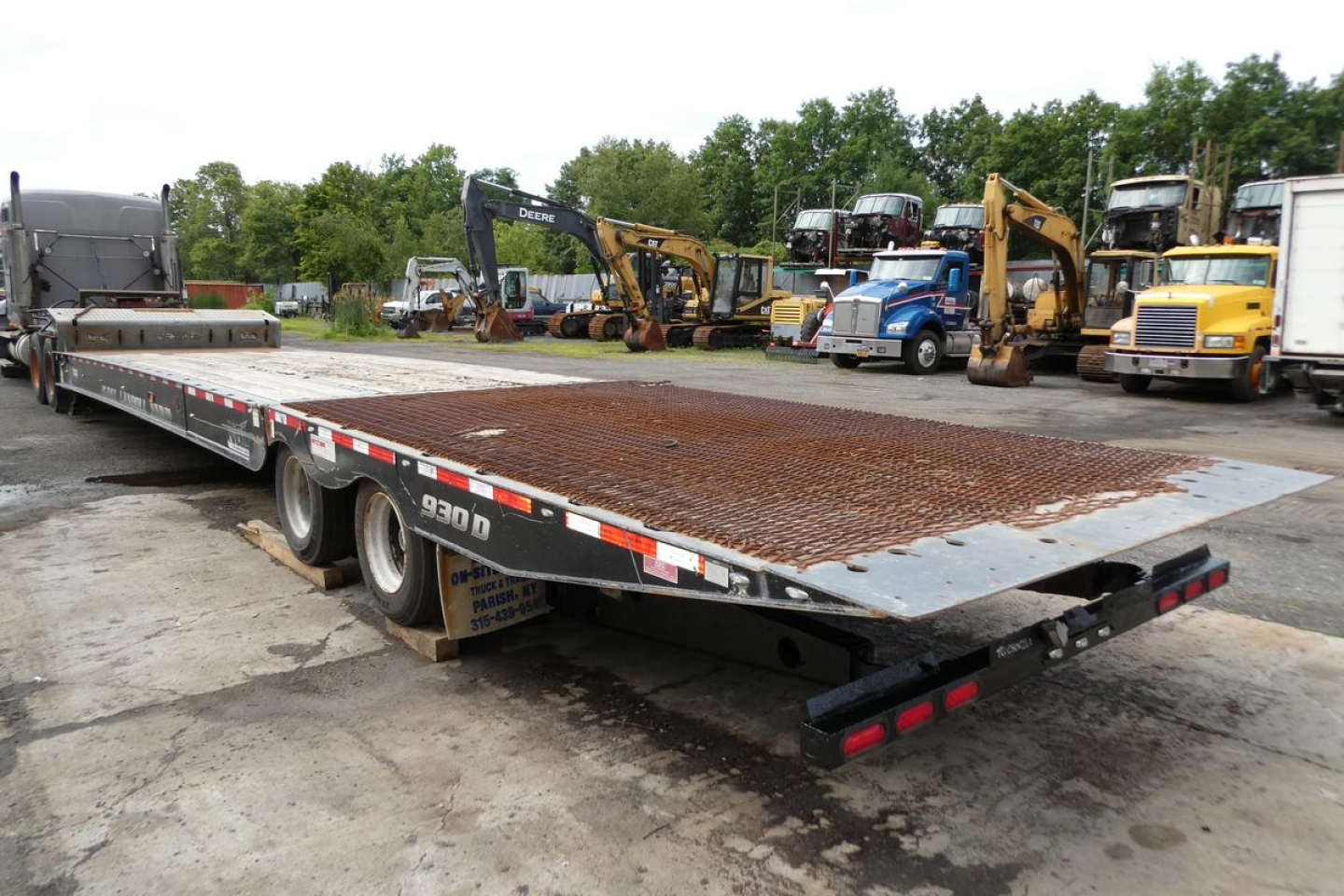 Flatbed trailer with a grated steel deck and triple-axle configuration, viewed from the rear-left. Industrial yard with excavators and trucks in the background.