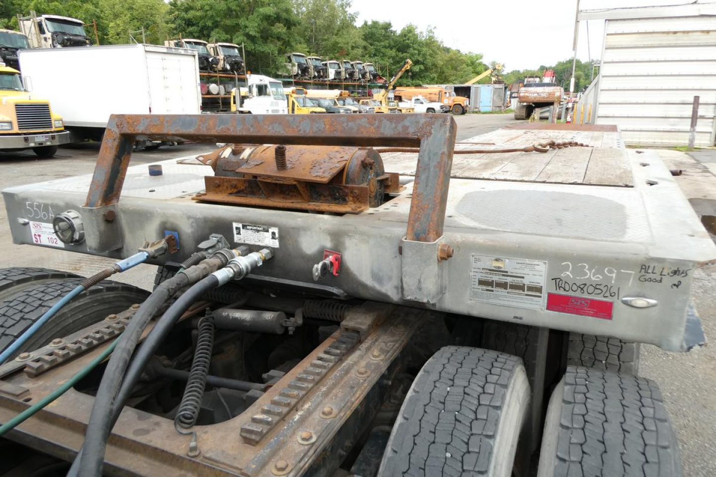 Trailer rear deck with hydraulic coupler and dual wheels, metal frame rails, and control levers; weathered gray surface with handwritten tags and red tag on right side.