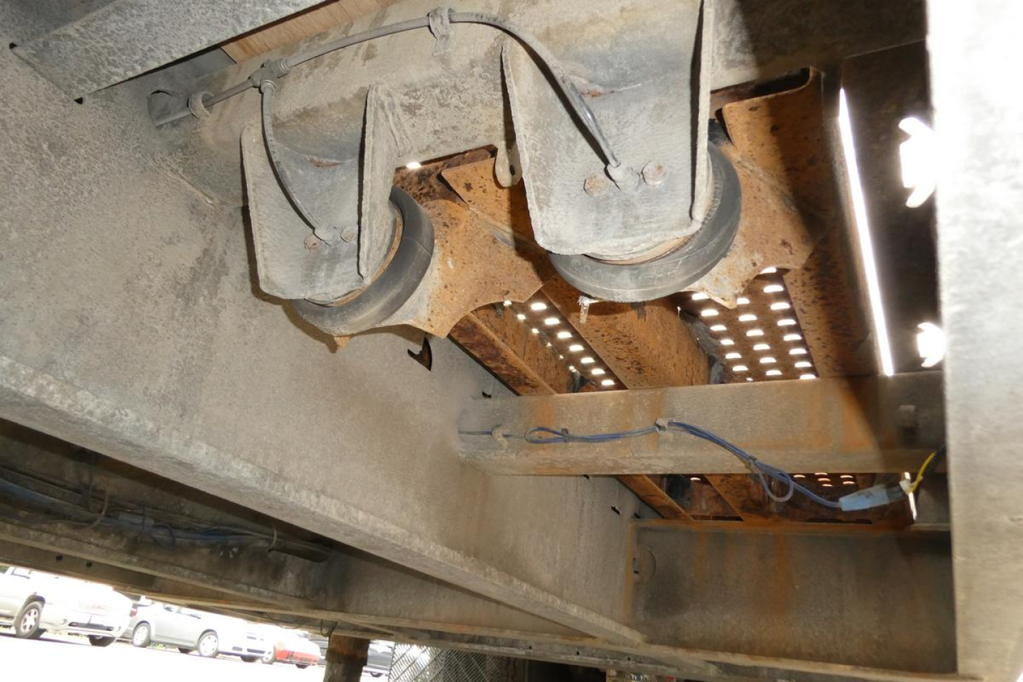 Trailer underside showing two axles with wheels and suspension, rusted frame, hoses, and perforated metal platform; no legible text present.