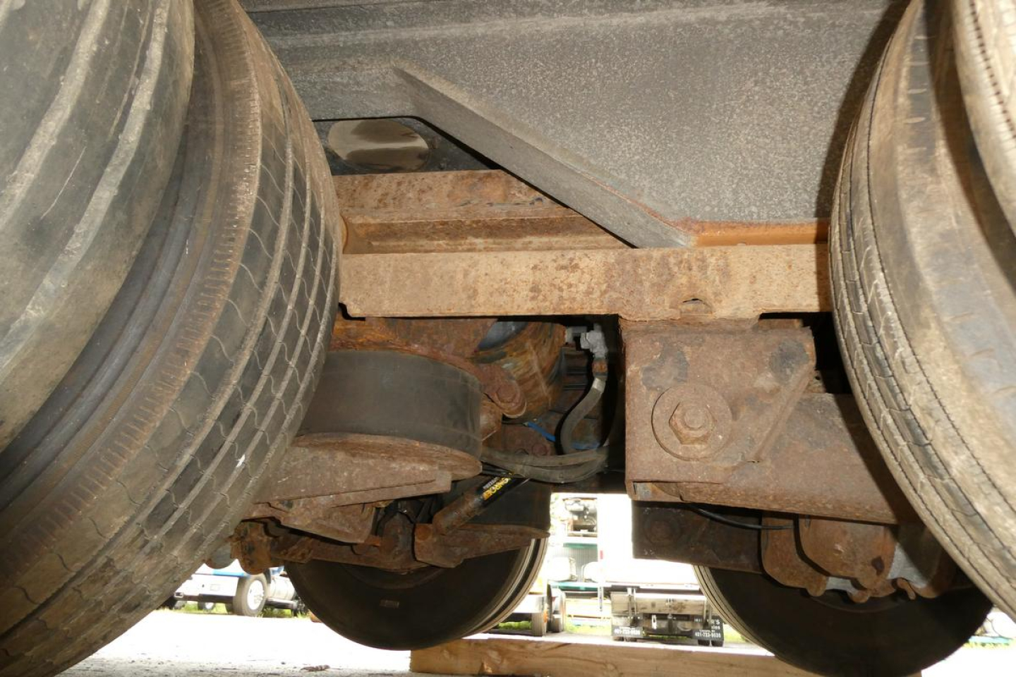 Trailer undercarriage showing dual rear axles with rusted steel frame and leaf springs; tires visible and some mechanical links near center