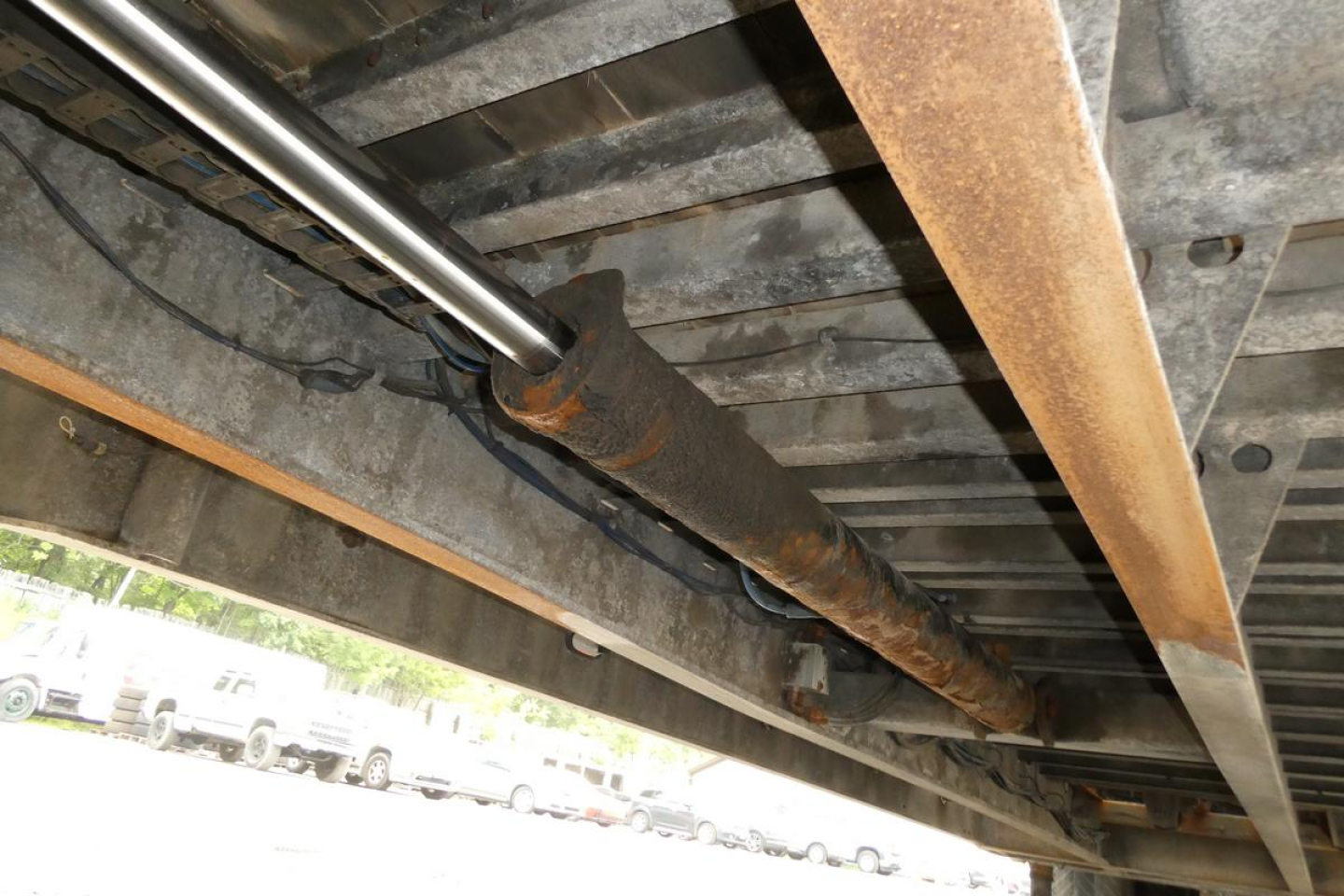 Trailer undercarriage view showing a hydraulic cylinder/prop and steel framing with rust-colored pipe; no legible model labels visible in image.