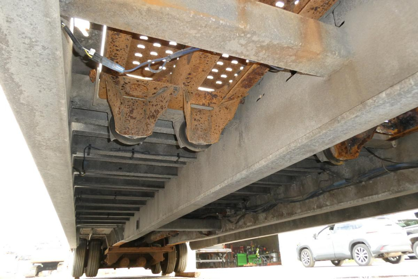 Vehicle trailer undercarriage showing steel I-beam frame and suspension components, with rust and hydraulic lines visible.