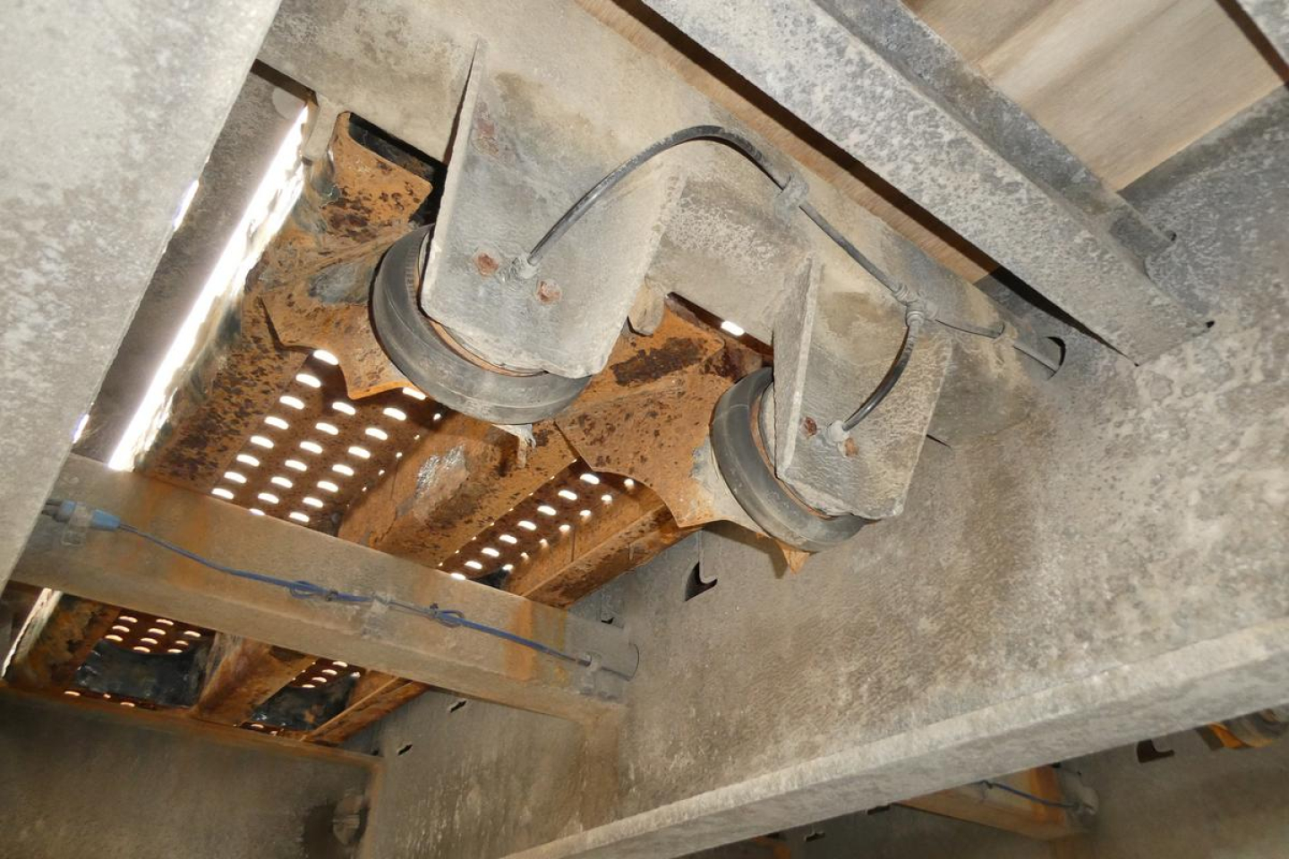 Trailer undercarriage showing dual axles with leaf spring suspension and rust-prone metal frame; visible wiring harness and underside components.
