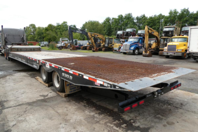 Flatbed trailer with a grated steel deck and triple-axle configuration, viewed from the rear-left. Industrial yard with excavators and trucks in the background.