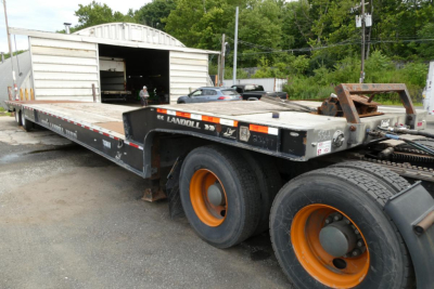 Landoll 930D NGL trailer with dual rear axles and orange rims, side rails showing branding, in a lot with a storage building in background.