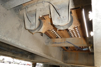 Trailer underside showing two axles with wheels and suspension, rusted frame, hoses, and perforated metal platform; no legible text present.