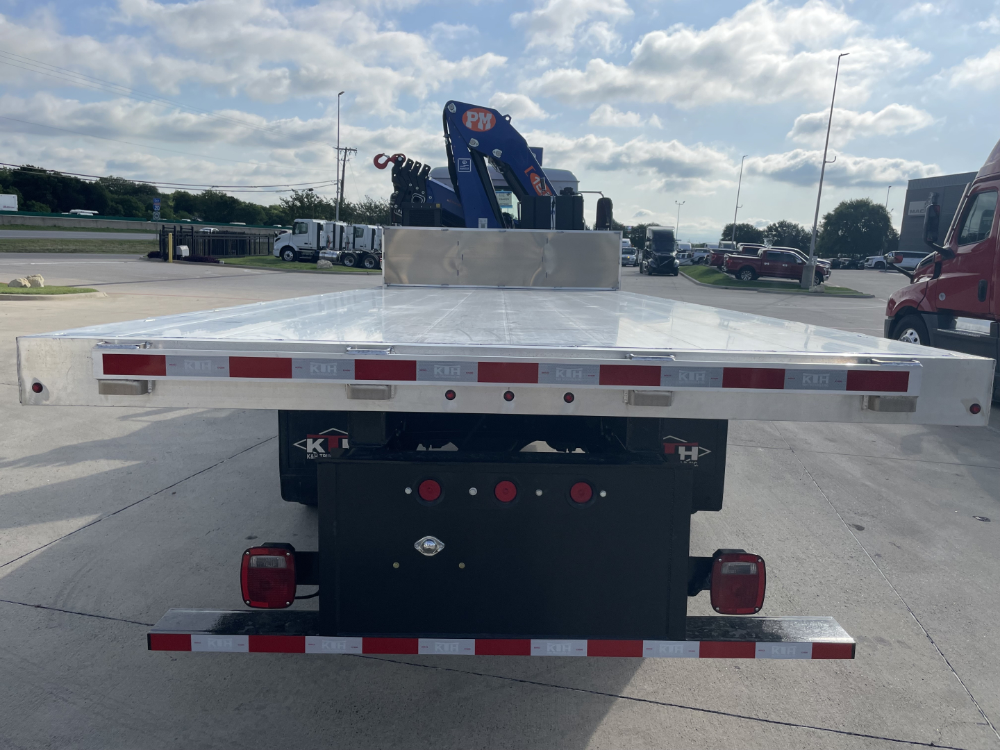 Flatbed truck bed with rear view showing reflective safety stripes and taillights, Mack MD7 boom truck in distance, chrome flat steel bed with toolbox area.