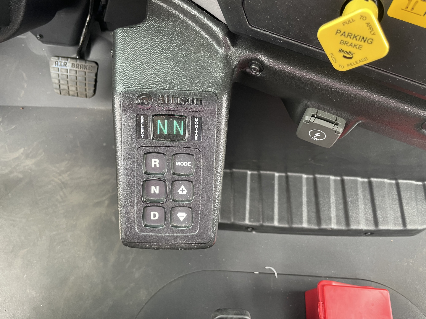 Allison transmission gear selector with NN engaged showing R N D with mode buttons, pedal area, yellow parking brake indicator, and grey dashboard near a red item.
