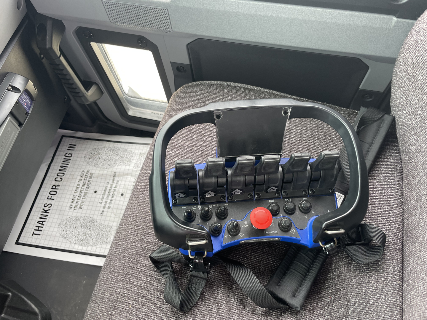 Interior of Mack MD7 cab showing cockpit joystick-like control panel with multiple blue levers, red emergency stop, black belt, and side window; light gray seats and dashboard edge visible.
