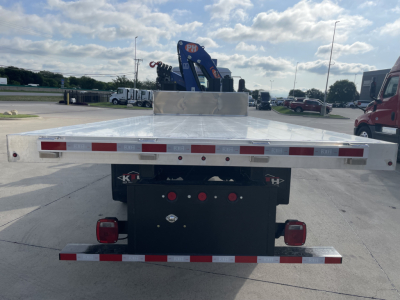 Flatbed truck bed with rear view showing reflective safety stripes and taillights, Mack MD7 boom truck in distance, chrome flat steel bed with toolbox area.