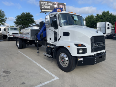 White Mack MD7 boom truck in parking lot, side view, with blue hydraulic crane arm and stabilizers extended. Visible text on truck includes MACK logo on grille and side mirrors; billboard in background partially visible.