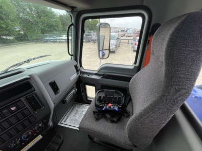 Truck cab interior of a 2025 Mack MD7 showing steering wheel, dashboard, and driver seat with placard on seat. Side mirror visible through window.