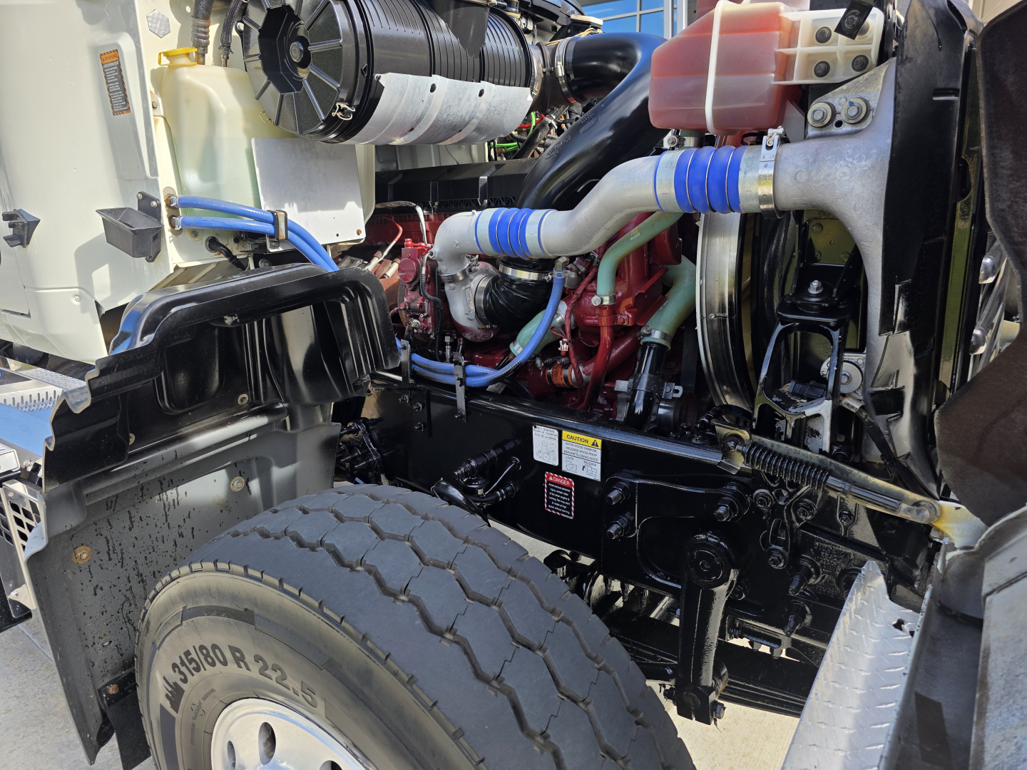Truck engine bay with large front tire visible and various hoses, hoses with blue cuffs, red engine block, and black chassis; visible coolant reservoir; part of Mack Granite cab-front area.