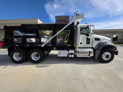 Dump truck side view showing Mack GRANITE with black dump body, chrome fuel tanks, and heavy-duty frame. Visible text includes MHC on building sign in background.