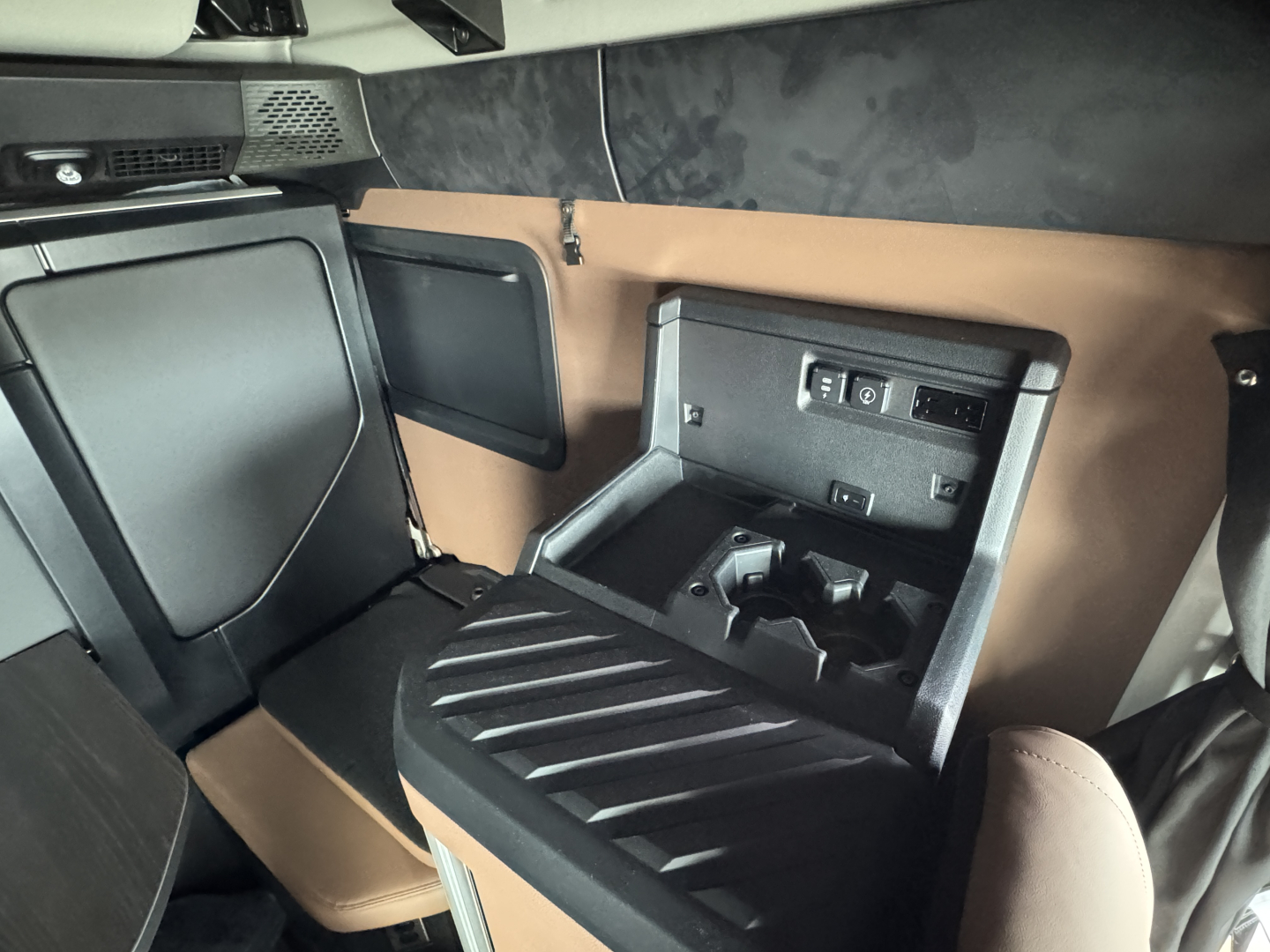 Truck cab interior showing storage console and cup holders in a Mack PR64T76H sleeper cab. Visible components include a center console with a recessed area and electrical outlets, brown and black interior panels, and a small overhead shelf.