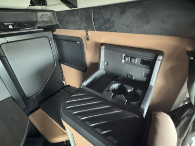 Truck cab interior showing storage console and cup holders in a Mack PR64T76H sleeper cab. Visible components include a center console with a recessed area and electrical outlets, brown and black interior panels, and a small overhead shelf.