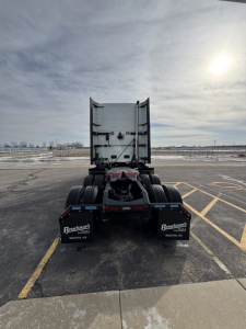 Rear view of a Mack PR64T76H truck tractor chassis in an empty lot; visible mud flaps show Buckner truck logos; no cab, fifth wheel area present.