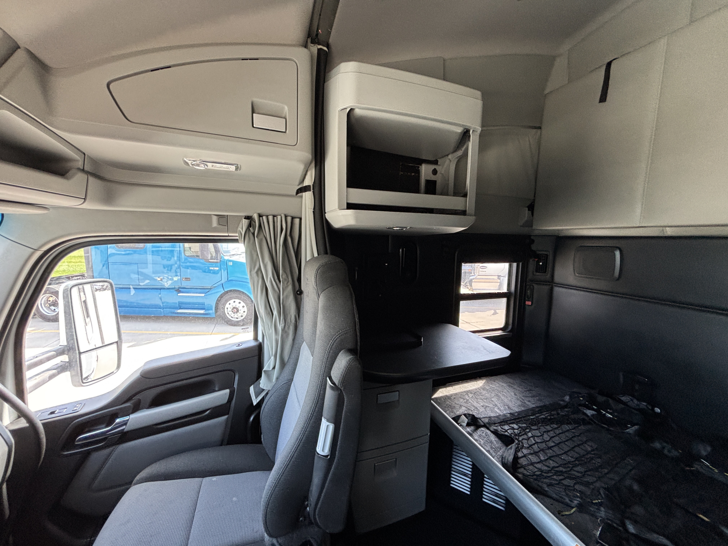 Cab interior of a 2022 Kenworth T680 with sleeper area, bunk, overhead storage, and window visible; dashboard to left, grey upholstery.