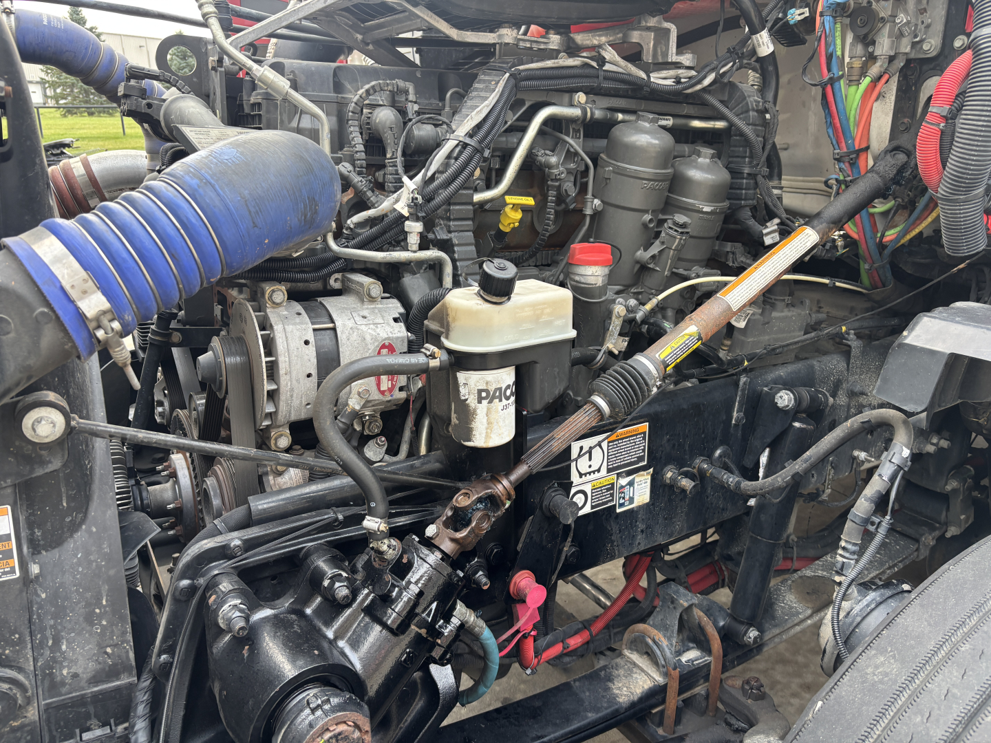 Kenworth T680 engine bay showing air intake, hoses, and various reservoirs; visible components include coolant reservoir, power steering reservoir, filters, and wiring harnesses.