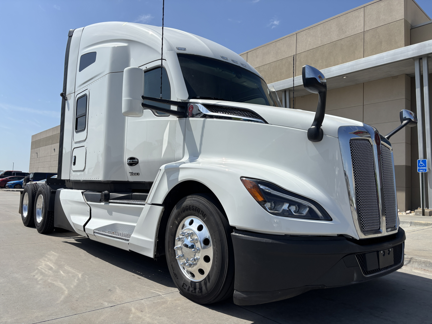 Kenworth T680 tractor-trailer in white, front-left view showing aerodynamic cab, large grille, dual side mirrors, and connected sleeper cabin.