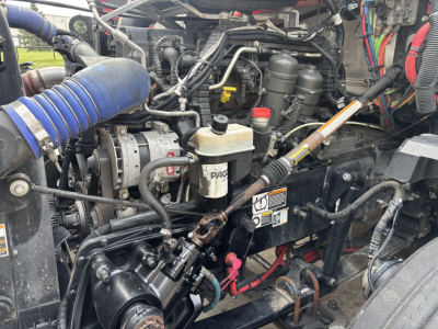 Kenworth T680 engine bay showing air intake, hoses, and various reservoirs; visible components include coolant reservoir, power steering reservoir, filters, and wiring harnesses.