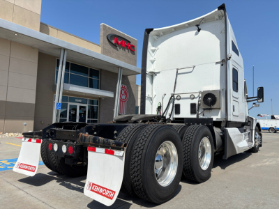 Kenworth T680 tractor with sleeper, rear view showing dual rear axles and fifth wheel, fuel tanks, and mud flaps in front of dealership.