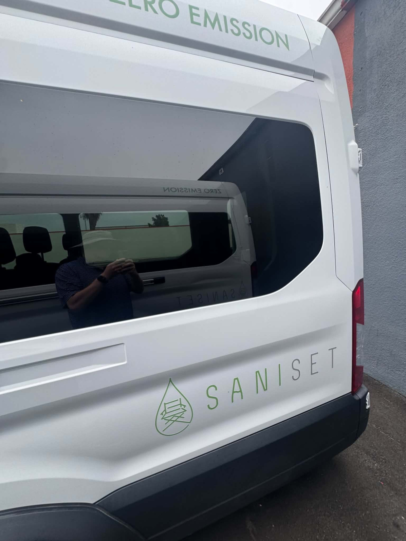 Ford Transit van rear side showing green zero-emission logo and SANISET branding on white exterior; rear window with passenger seating visible inside; partial reflection of person in glass.