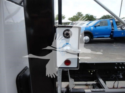 Truck equipment panel with circular gauge and red indicator on a roll-off truck bed, viewed from side with blue pickup in background.