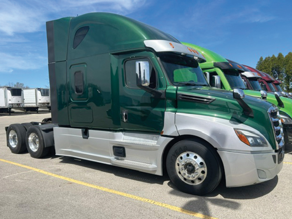 Truck cab in a row of Freightliner Cascadia models showing green and silver exterior, with sleeper cab and multiple axles visible.
