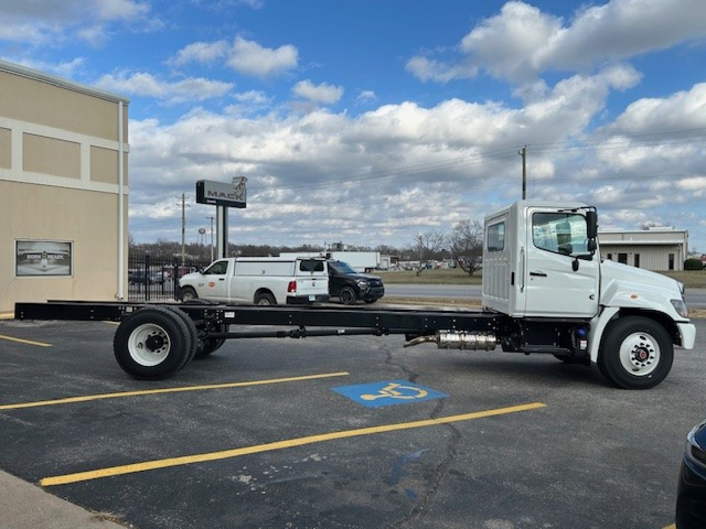 Hino L6 cab chassis truck in a parking lot, side view showing long chassis with no cargo bed, white cab, black undercarriage, and spare wheels visible.
