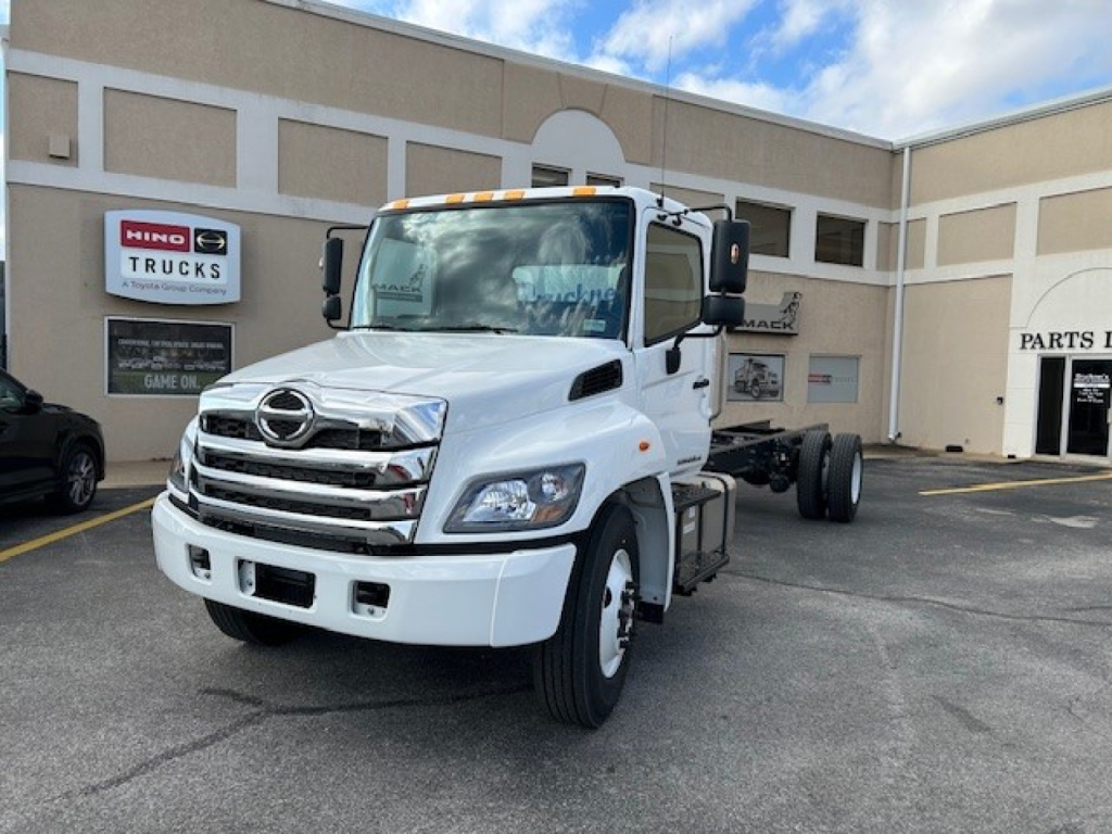 2025 Hino L6 cab and chassis truck in white, front-left view, with Hino Trucks sign in background; chrome grille, rectangular headlights, dual rear wheels, side mirrors, orange clearance lights on roof.