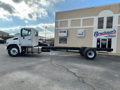 Side view of a white 2025 Hino L6 cab and chassis truck in a parking lot showing the long bare frame with dual rear wheels and building in background.