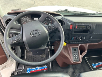 Cab and chassis dashboard view of 2025 Hino L6 showing steering wheel, instrument cluster, center console with controls, and brown gear area.