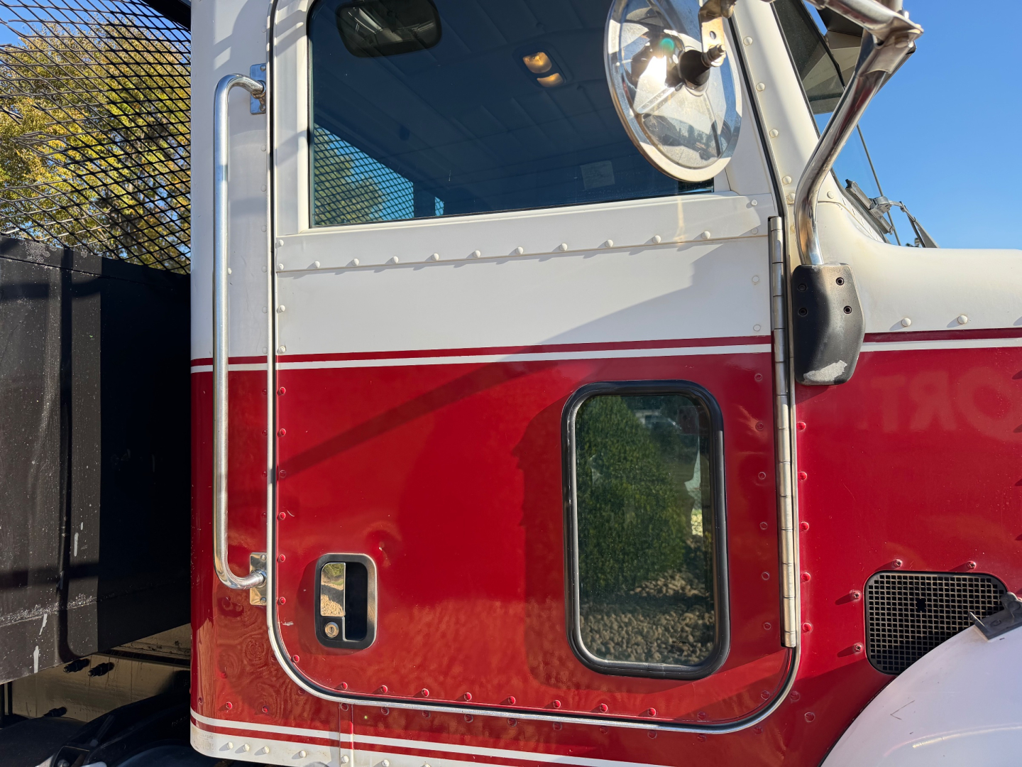 Peterbilt truck cab front left door area with red and white two-tone paint, chrome side mirror, and rectangular window; part of flatbed truck visible.