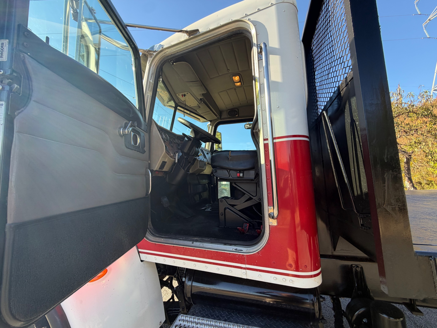 Peterbilt 335 cab interior view from open driver door; dash, steering wheel, seat, and door panel visible; red lower cab paint with white trim; flatbed truck area in background