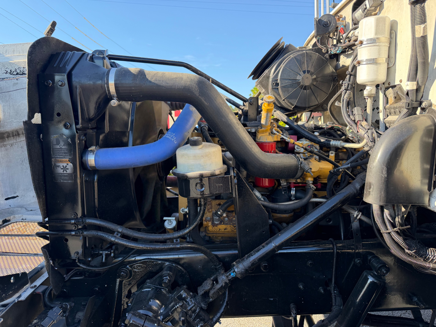 Peterbilt engine bay showing large cooling fan, blue radiator hose, black intake hose, yellow engine block, and various hydraulic lines. Visible components include radiator, coolant reservoir, and engine accessories.