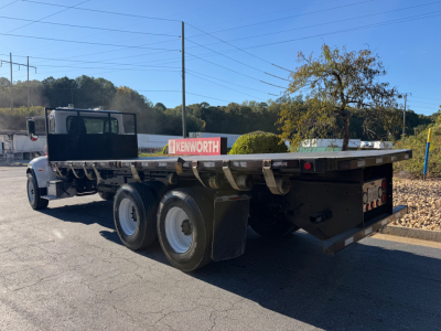Peterbilt 335 flatbed truck rear view showing dual rear axles and flatbed with a Kenworth sign in background, VIN and stock number not visible on image