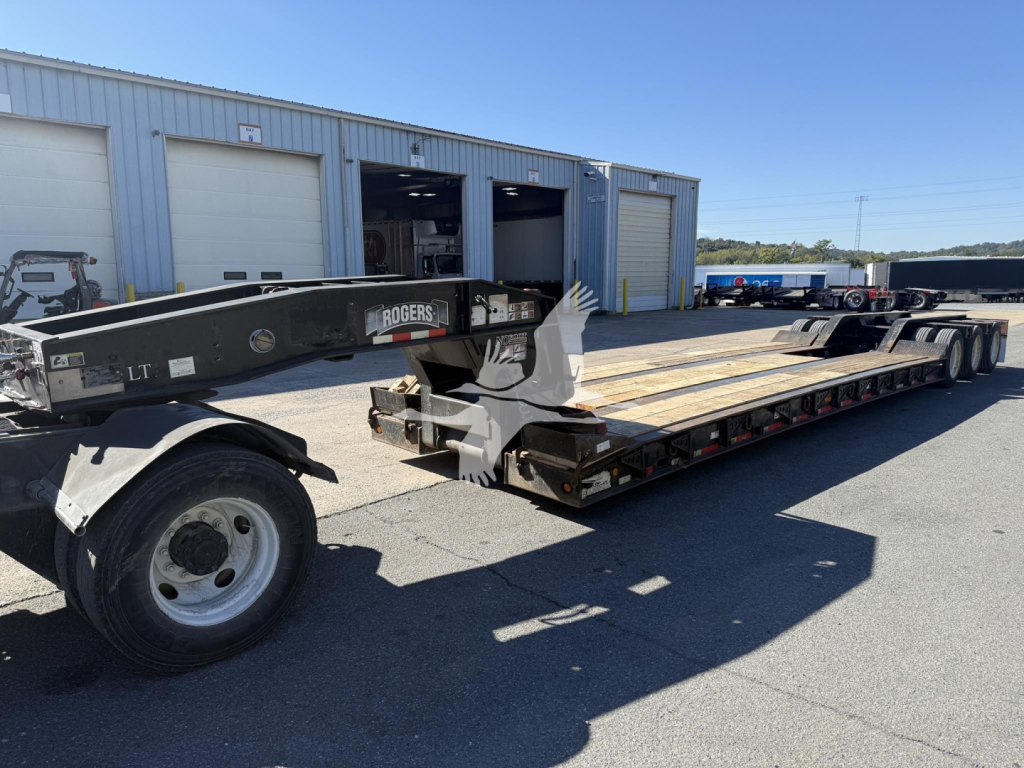 Trailer with Rogers 50-Ton lowboy showing long flatbed, dual rear axles, and front gooseneck with Rogers branding; light gray metal building in background, clear day.