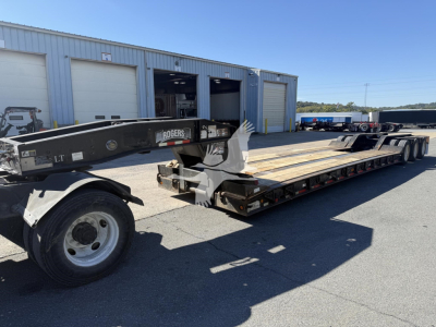 Trailer with Rogers 50-Ton lowboy showing long flatbed, dual rear axles, and front gooseneck with Rogers branding; light gray metal building in background, clear day.