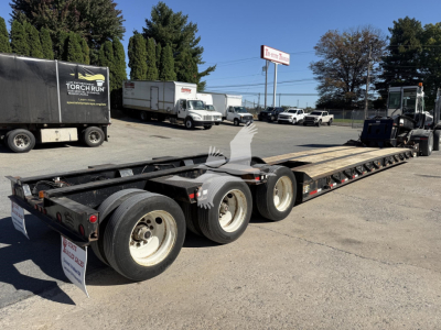 Long lowboy trailer with multiple axles, Rogers 50-ton model. Visible text includes company signs and license details on the trailer and nearby vehicles.