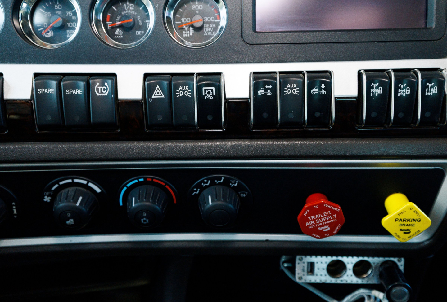 Cockpit dashboard of a Kenworth truck showing gauges, switches, and control knobs with circular gauges and warning/toggle buttons.