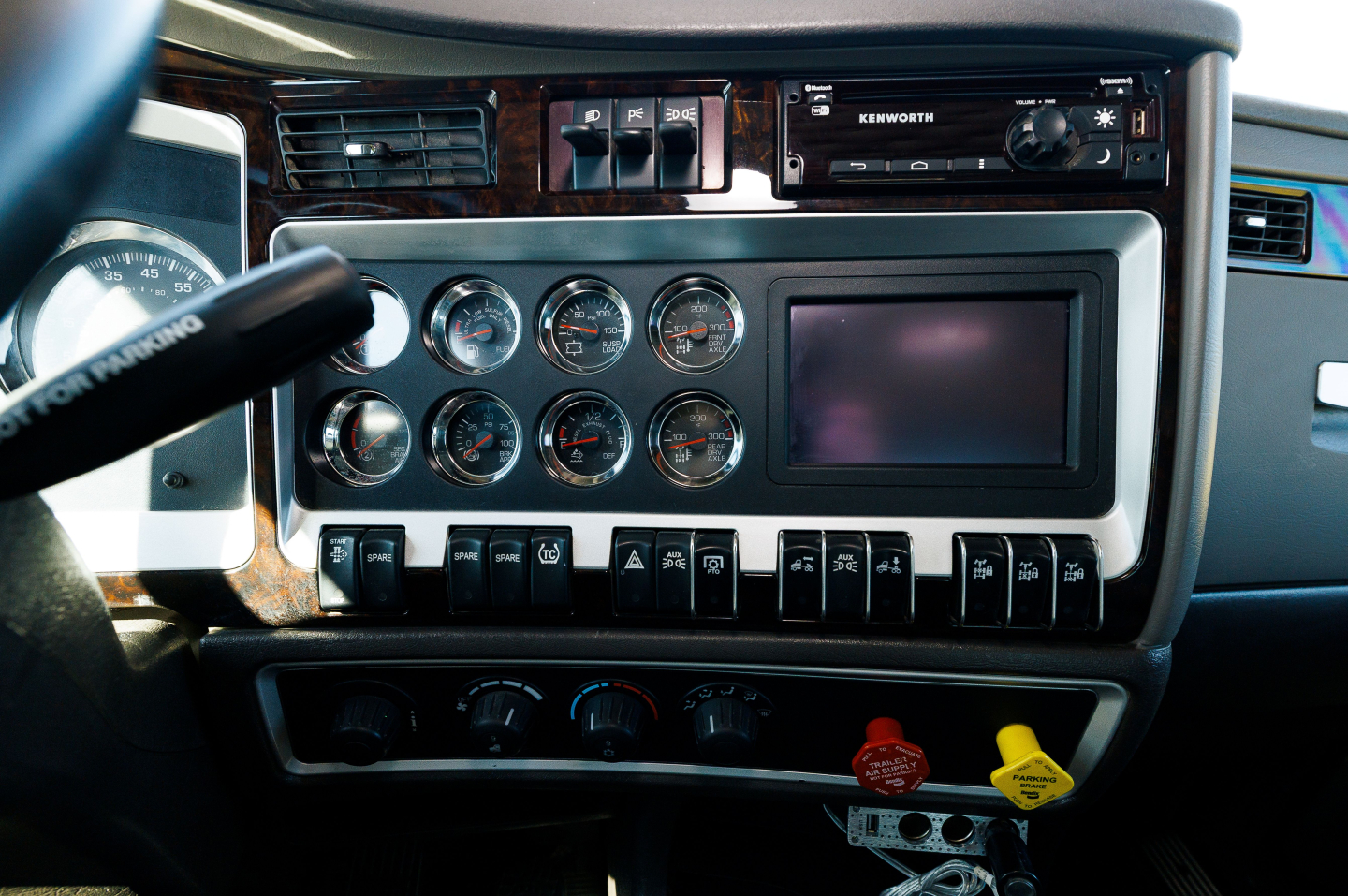 Dashboard view of a Kenworth truck cab showing multiple round gauges, a touchscreen, switches, and ignition area.